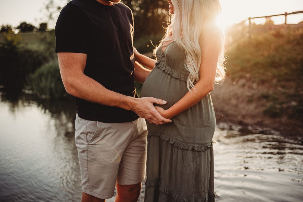 a man and woman standing facing each other in a river. Their hands are on he growing baby bump