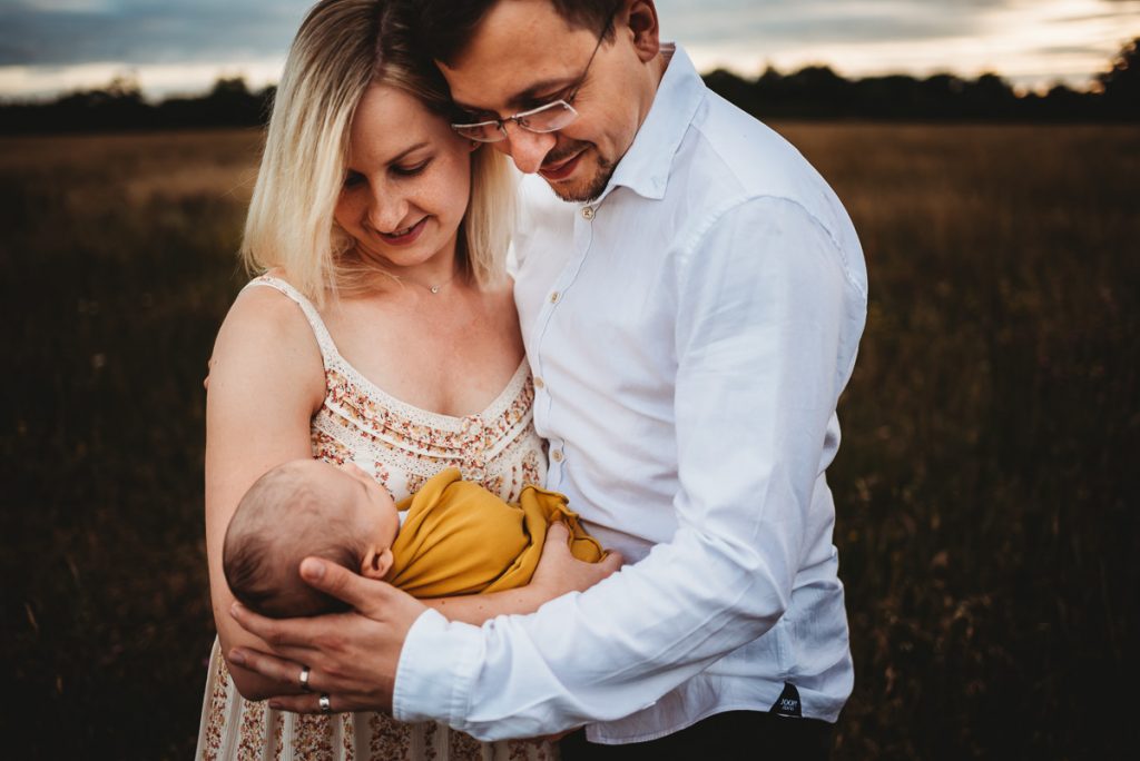 mum and dad standing close together with their newborn baby at sunset