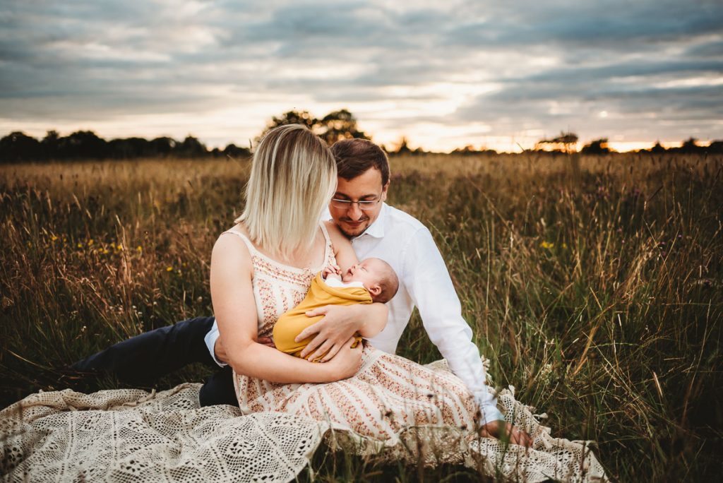 mother and father cuddling their newborn baby in a field of long grass at sunset