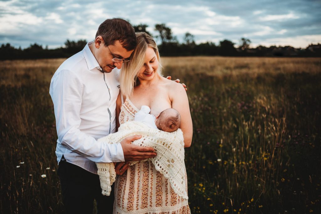 mother breastfeeding her baby in a filed with her partner standing next to her. Outdoor newborn photography during sunset