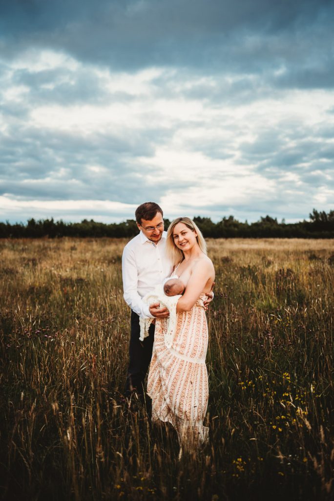 mother and father standing in a field with their newborn baby. Mother is breastfeeding the baby