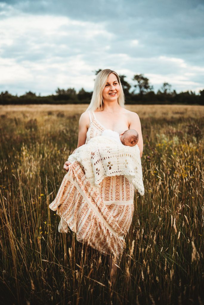 woman wearing a long dress stands in a field breast-feeding her newborn baby