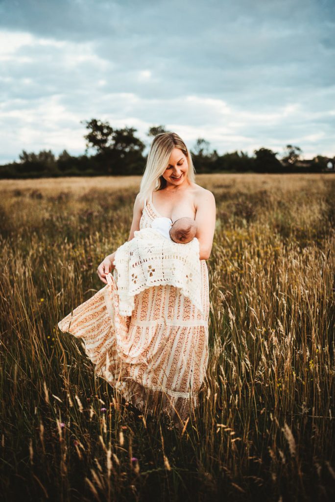 woman wearing a long dress stands in a field breast-feeding her newborn baby