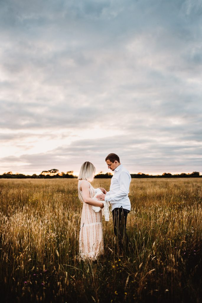 mum and dad standing in a filed with their newborn baby. outdoor newborn photography during sunset