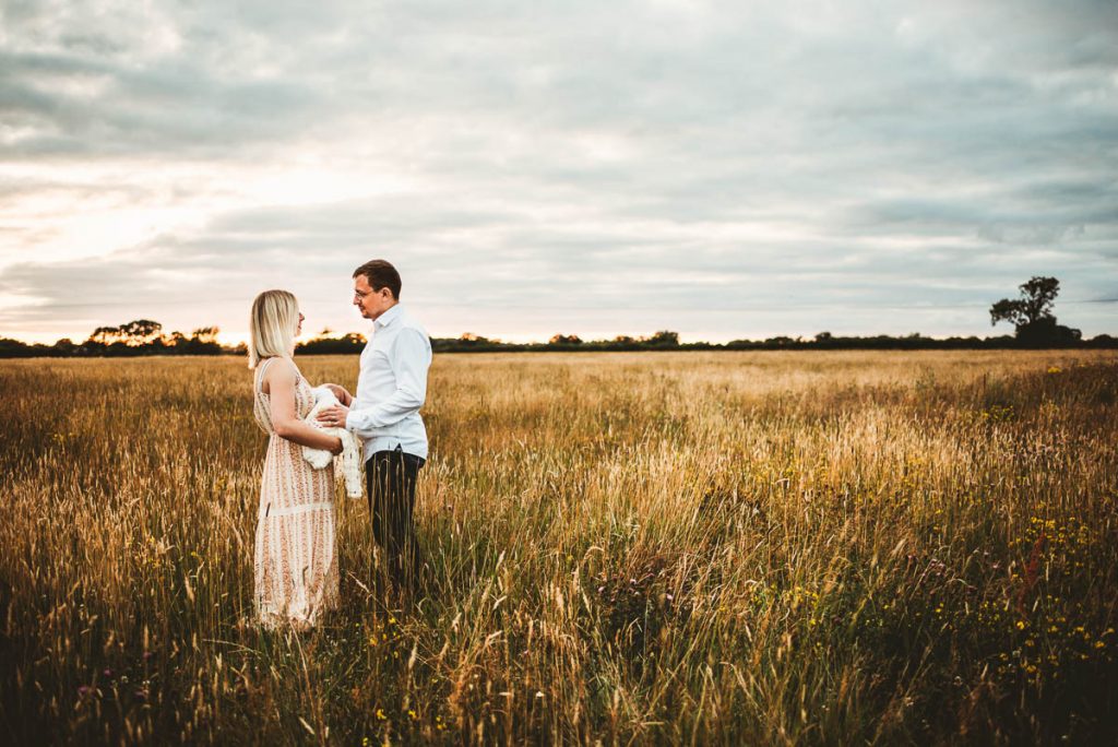 mum and dad facing each other in a golden field. Their newborn baby is between them