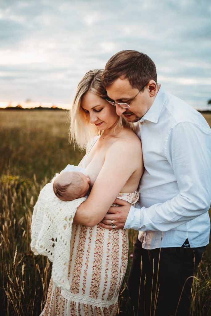 a mother breastfeeding her newborn baby outside whilst her husband looks over her shoulder at the newborn baby