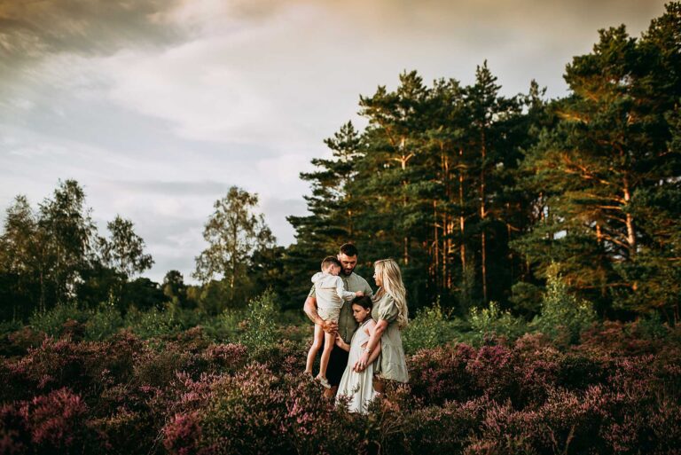 family of four standing in purple heather with the sunset and woodland trees behind them