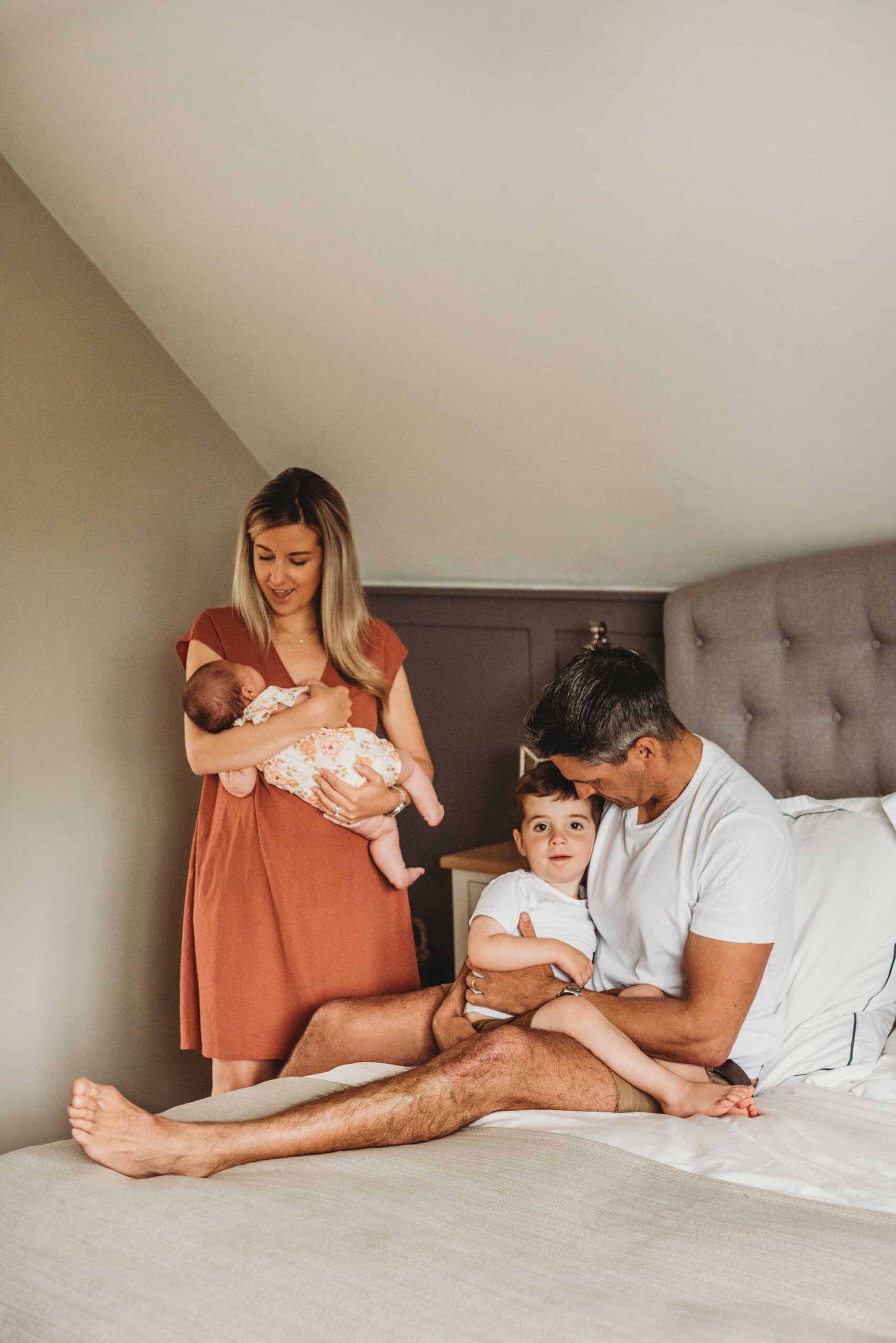 dad sitting on the bed with little boy whilst mum stands holding newborn baby