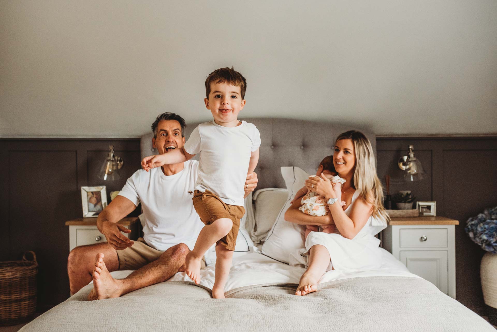 little boy jumping on the bed with his parents and baby sister behind him