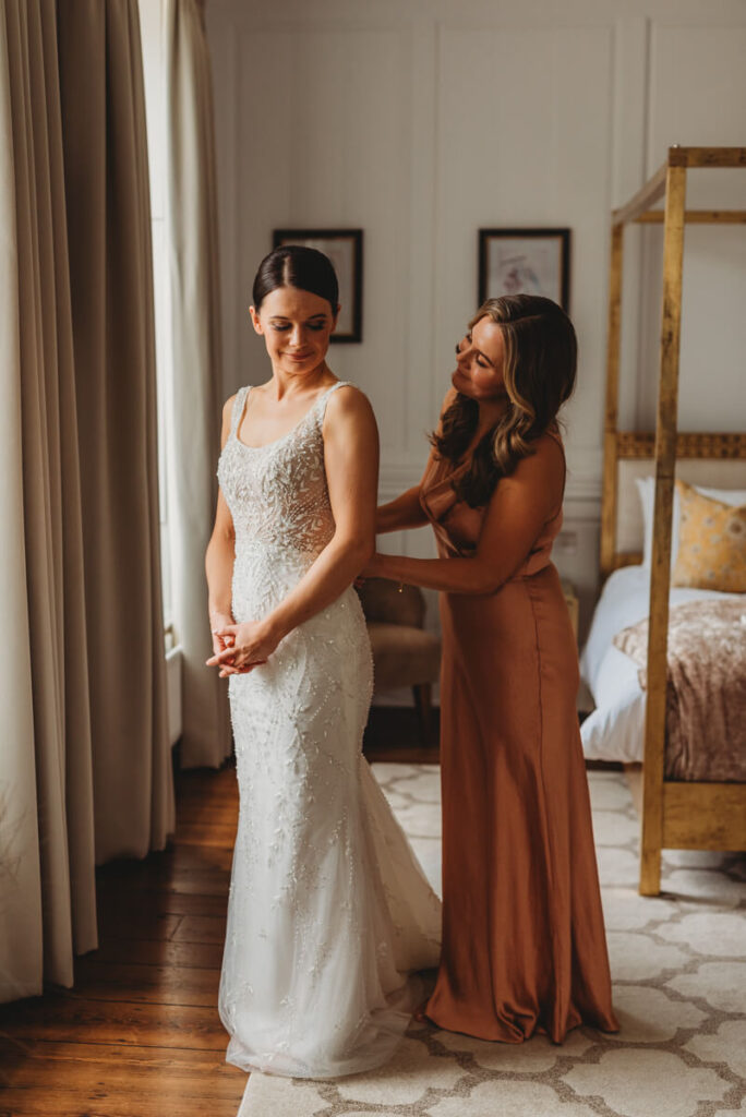 bridesmaid helping the bride to fasten her dress by Wedding Photographer in Wiltshire, the Cotswolds, Gloucestershire and beyond.