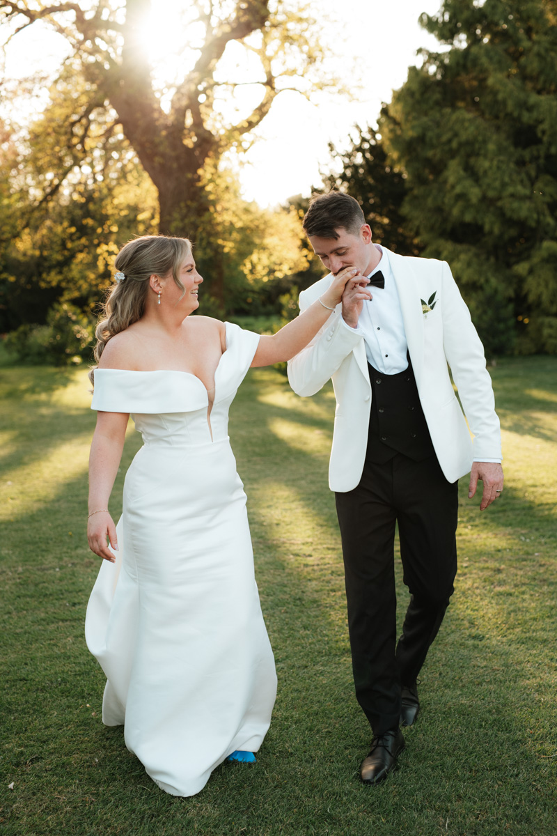 bride and groom walking through the gardens at Grittleton house in Wiltshire. Groom is kissing his bride on the hand as they walk