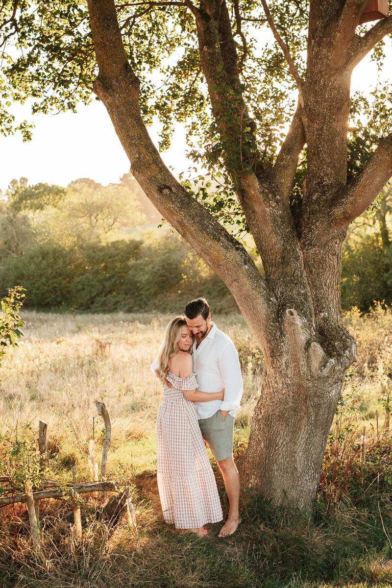 engaged couple standing under a tree during sunset 