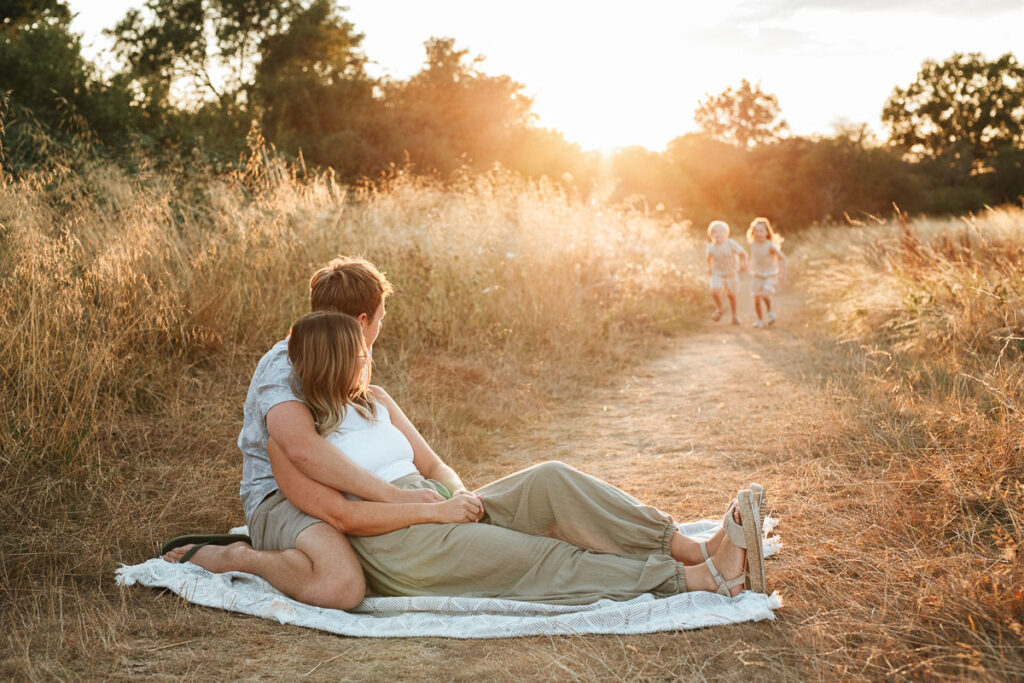 parents sitting together on a blanket watching their children run towards them during their family photography session with Rachel Jane