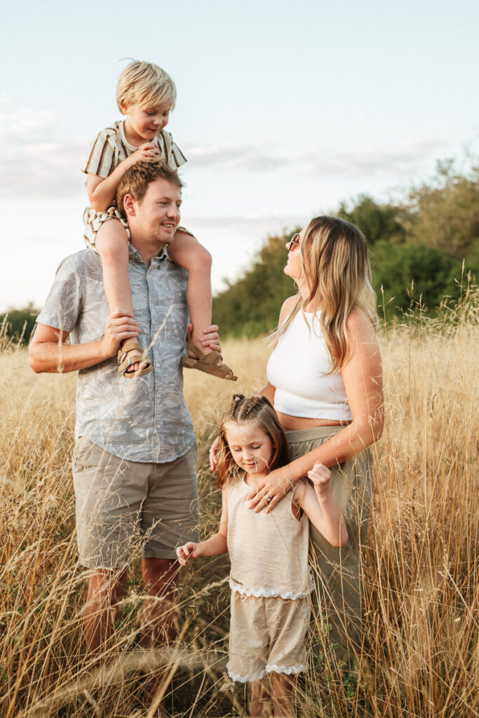parents and children standing together in golden grass. Dad has one child on his shoulders. Captured by Wiltshire family photographer Rachel Jane