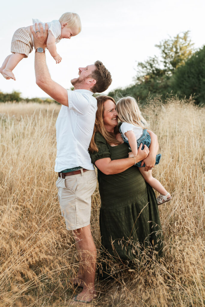 parents standing back to back holding their two young children as they laugh and cuddle. Sunset family photography by Wiltshire photographer Rachel Jane