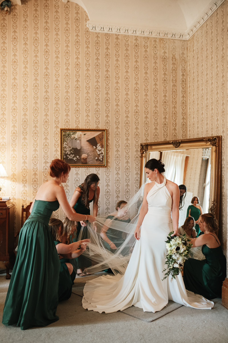Wiltshire wedding photographer bride getting ready surrounded by her bridesmaids as they all help arrange her dress and veil